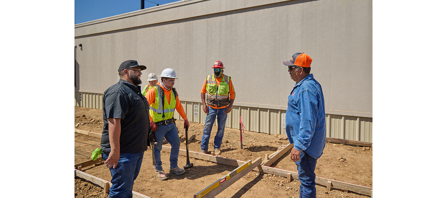 Five construction workers stand at a job site discussing plans near a foundation layout.
